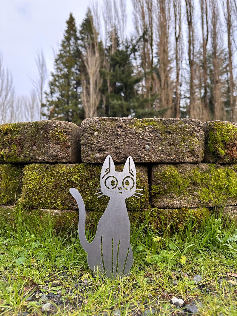 May include: A silver metal cat silhouette with large eyes and whiskers sits in front of a moss-covered brick wall. The cat is in a seated position with its tail curved. The background includes green grass and trees.
