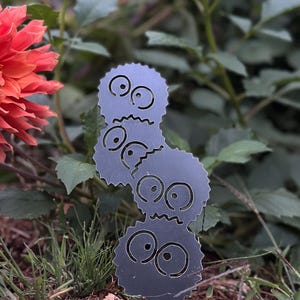 K&ouml;nnte beinhalten: Ein Gartenstecker aus dunkelgrauem Metall mit drei gestapelten Cartoon-Gesichtern mit gro&szlig;en Augen und gezackten M&uuml;ndern. Der Stecker befindet sich in einem Garten mit gr&uuml;nem Laub und einer roten Blume.