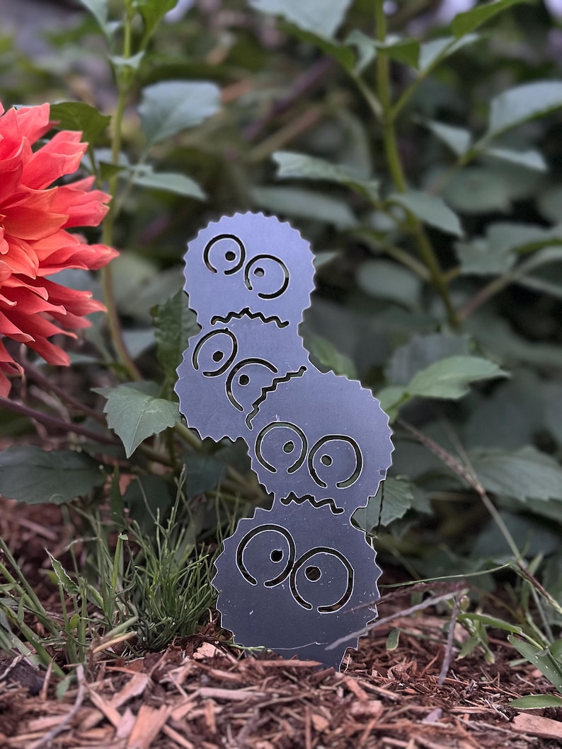 K&ouml;nnte beinhalten: Ein Gartenstecker aus dunkelgrauem Metall mit drei gestapelten Cartoon-Gesichtern mit gro&szlig;en Augen und gezackten M&uuml;ndern. Der Stecker befindet sich in einem Garten mit gr&uuml;nem Laub und einer roten Blume.