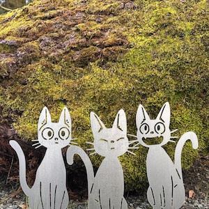 May include: Three metal cat yard art pieces with different facial expressions. The cats are silver and sit on a bed of small rocks in front of a moss-covered log. The cats have cut-out details.