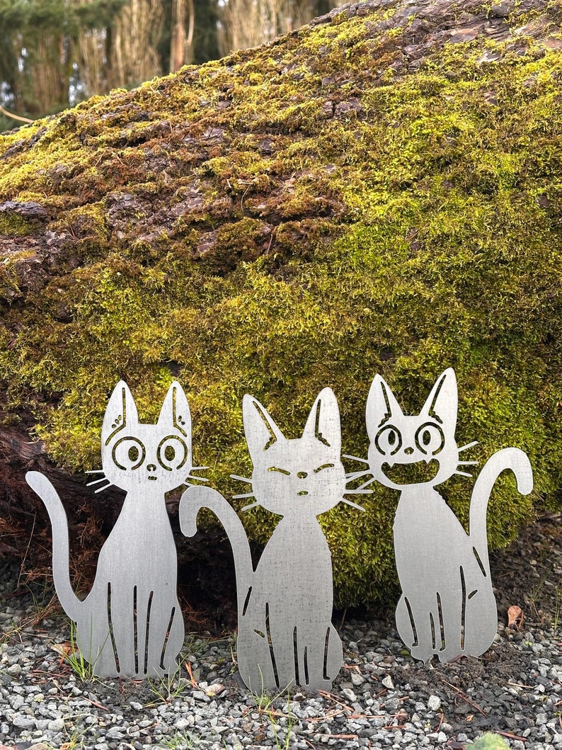 May include: Three metal cat yard art pieces with different facial expressions. The cats are silver and sit on a bed of small rocks in front of a moss-covered log. The cats have cut-out details.