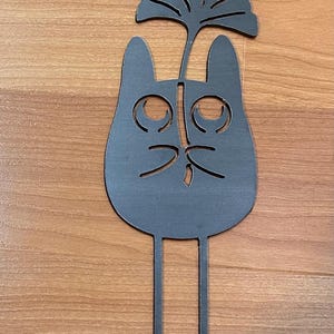 May include: A dark gray metal cat-shaped garden stake with a stylized leaf design above its head. The cat has large, round eyes and a simple nose and mouth. Two thin stakes extend from the bottom for insertion into the ground.