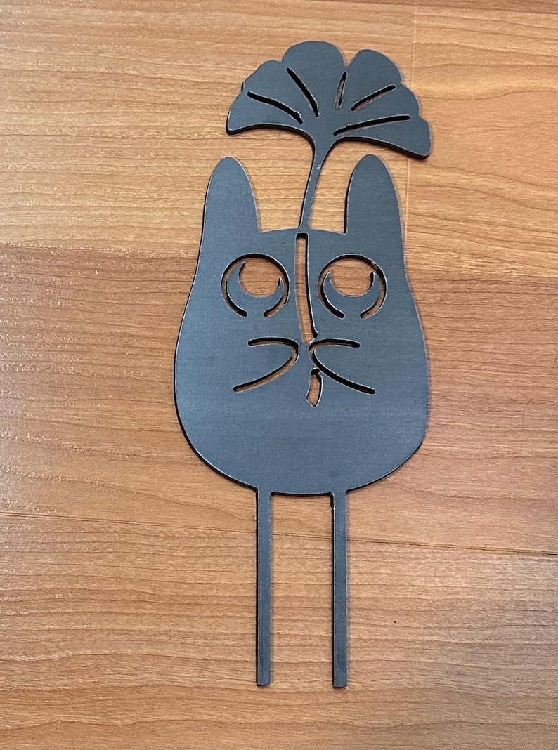 May include: A dark gray metal cat-shaped garden stake with a stylized leaf design above its head. The cat has large, round eyes and a simple nose and mouth. Two thin stakes extend from the bottom for insertion into the ground.