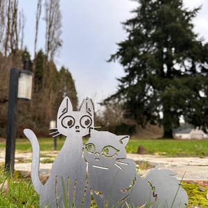 May include: Two metal cat sculptures, one upright and one seated, are displayed in a grassy area. The cats are cut from a silver-colored metal and have detailed facial features. The background includes trees and a cloudy sky.