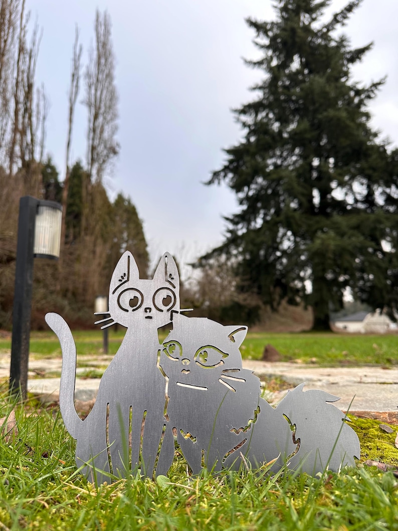 May include: Two metal cat sculptures, one upright and one seated, are displayed in a grassy area. The cats are cut from a silver-colored metal and have detailed facial features. The background includes trees and a cloudy sky.
