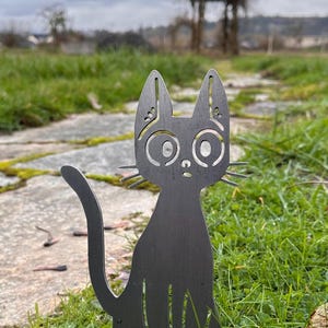 May include: A black metal cat garden stake with large, round eyes and pointed ears. The cat is sitting upright in a grassy area with a stone path. The cat's tail curves upwards. The background includes a blurred view of a garden and cloudy sky.