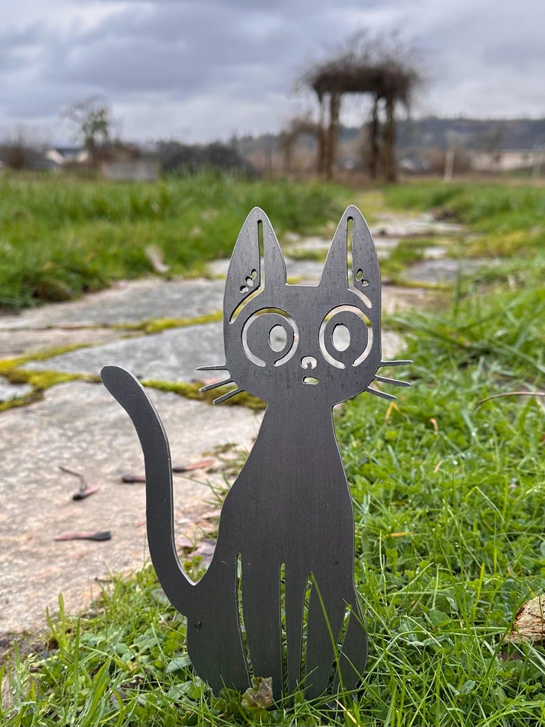 May include: A black metal cat garden stake with large, round eyes and pointed ears. The cat is sitting upright in a grassy area with a stone path. The cat's tail curves upwards. The background includes a blurred view of a garden and cloudy sky.