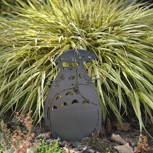 May include: A dark gray metal garden sculpture of a Totoro character holding an umbrella. The sculpture is placed in a garden bed with green and yellow ornamental grass in the background. The sculpture is approximately 12 inches tall.