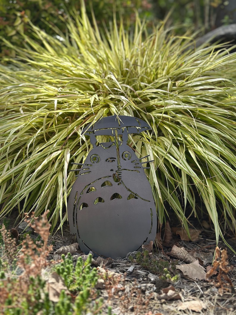 May include: A dark gray metal garden sculpture of a Totoro character holding an umbrella. The sculpture is placed in a garden bed with green and yellow ornamental grass in the background. The sculpture is approximately 12 inches tall.