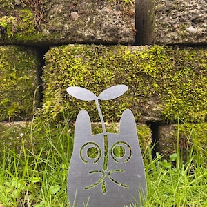 May include: A gray metal cat-shaped garden stake with a sprout growing from its head. The cat has large, round eyes and a smiling mouth. The stake is set in green grass, with moss-covered bricks in the background.