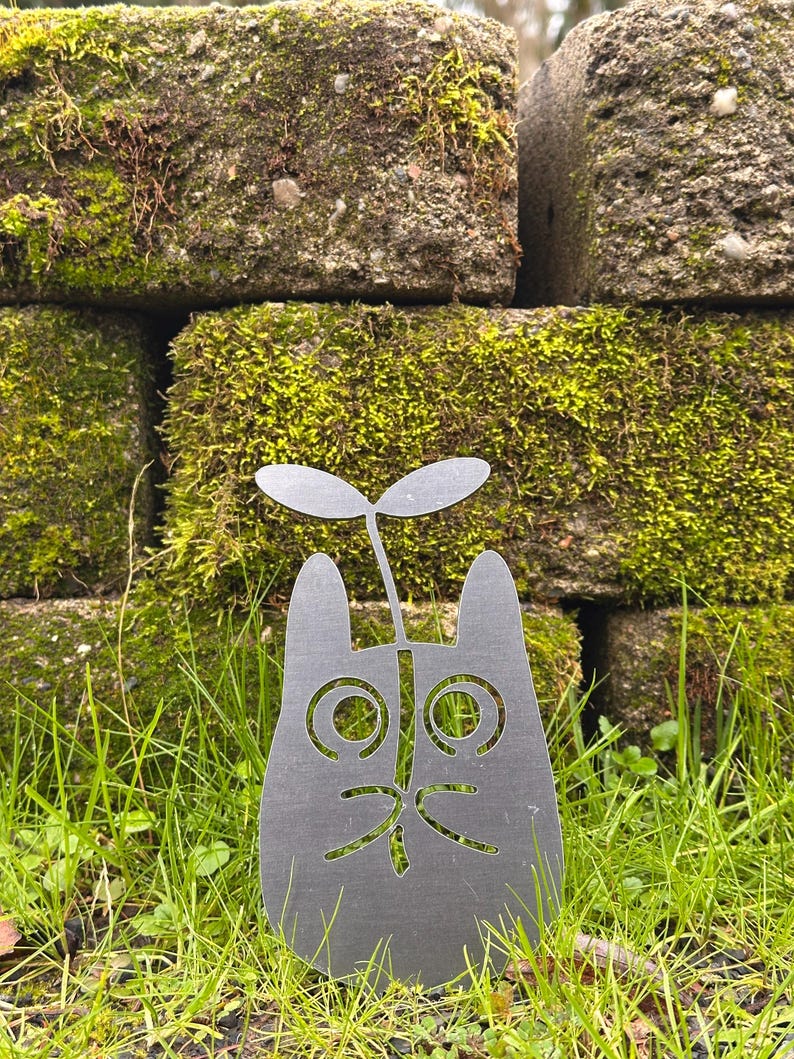 May include: A gray metal cat-shaped garden stake with a sprout growing from its head. The cat has large, round eyes and a smiling mouth. The stake is set in green grass, with moss-covered bricks in the background.