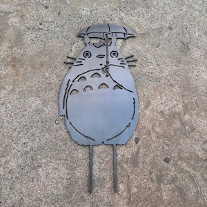 May include: A metal garden stake featuring a stylized depiction of Totoro holding an umbrella. The gray silhouette has cut-out details and two stakes for ground insertion. The design is based on the animated film.