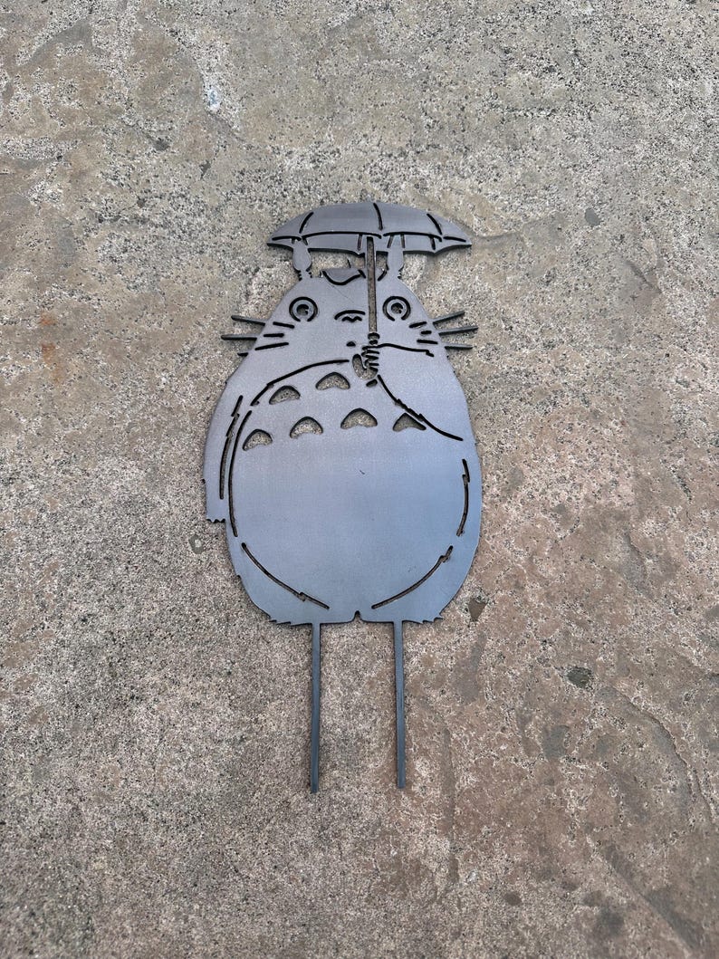 May include: A metal garden stake featuring a stylized depiction of Totoro holding an umbrella. The gray silhouette has cut-out details and two stakes for ground insertion. The design is based on the animated film.