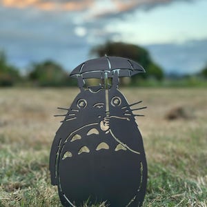 May include: A black metal sculpture of a cartoon character holding an umbrella. The character has a round body, pointed ears, and whiskers. The sculpture is placed in a grassy area with a blurred background of trees and a colorful sky.