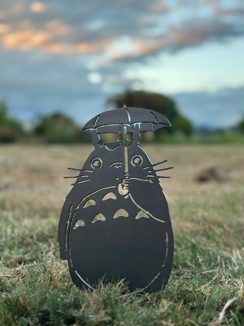 May include: A black metal sculpture of a cartoon character holding an umbrella. The character has a round body, pointed ears, and whiskers. The sculpture is placed in a grassy area with a blurred background of trees and a colorful sky.