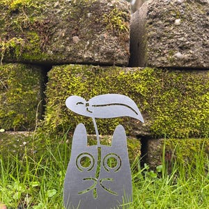 May include: A metal cat garden stake with a leaf accent. The cat has large, round eyes and a whimsical expression. The stake is set in green grass, with a mossy brick wall in the background.