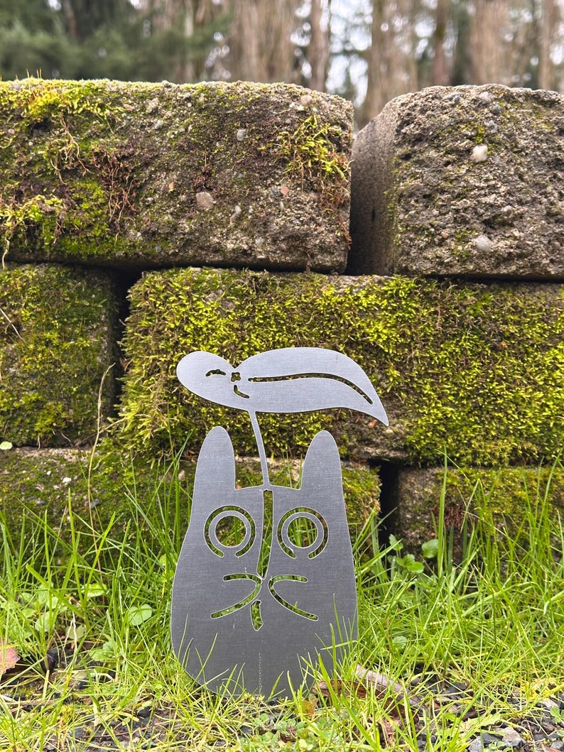 May include: A metal cat garden stake with a leaf accent. The cat has large, round eyes and a whimsical expression. The stake is set in green grass, with a mossy brick wall in the background.