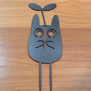 May include: A whimsical, dark gray metal cat-shaped garden stake. The cat has large, circular eyes, a small nose, and whiskers. Two straight legs extend downward, and a sprout with two leaves sits atop the cat's head. The stake is designed for outdoor use.
