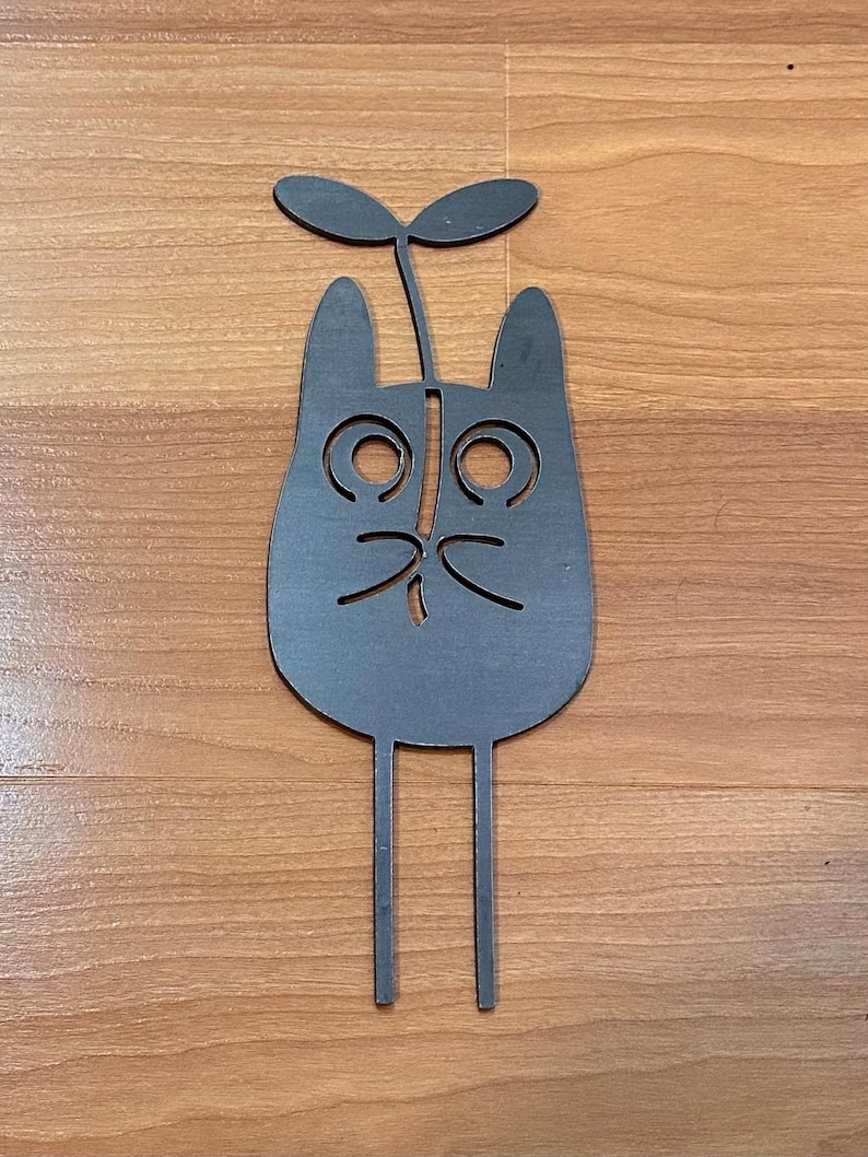 May include: A whimsical, dark gray metal cat-shaped garden stake. The cat has large, circular eyes, a small nose, and whiskers. Two straight legs extend downward, and a sprout with two leaves sits atop the cat's head. The stake is designed for outdoor use.