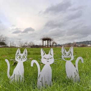 May include: Three metal cat yard art pieces with different facial expressions are placed in a grassy area. The cats are silver-colored and have cut-out designs. The background features a cloudy sky and a distant structure.