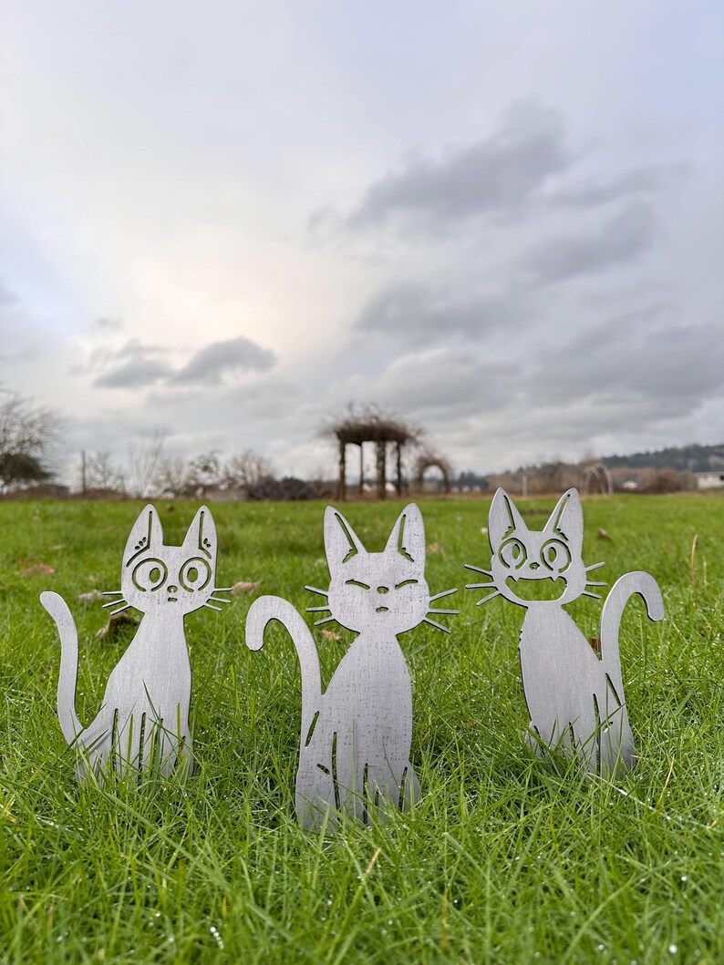 May include: Three metal cat yard art pieces with different facial expressions are placed in a grassy area. The cats are silver-colored and have cut-out designs. The background features a cloudy sky and a distant structure.