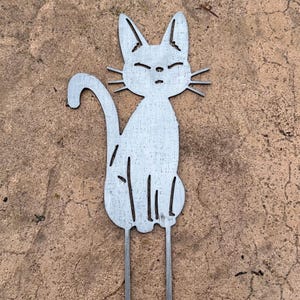 May include: A metal cat garden stake with a seated cat silhouette. The cat has pointed ears, closed eyes, and whiskers. The cat's tail curves upwards, and two metal rods extend from the bottom for insertion into the ground.