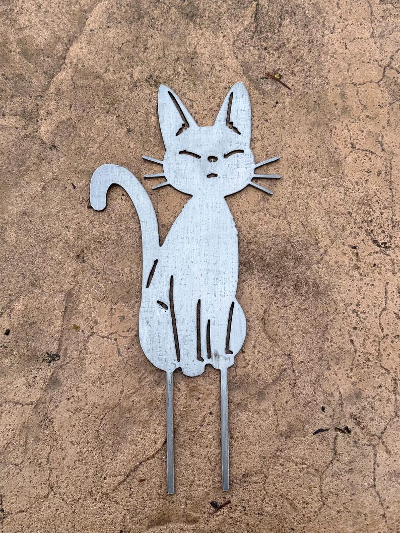 May include: A metal cat garden stake with a seated cat silhouette. The cat has pointed ears, closed eyes, and whiskers. The cat's tail curves upwards, and two metal rods extend from the bottom for insertion into the ground.