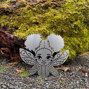 May include: A silver metal fairy with large, detailed wings and two circular puffs of hair. The fairy is resting on a bed of small, dark gray stones, with a mossy log in the background.