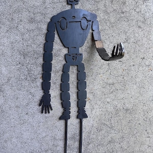 May include: A black metal robot figure with outstretched arms and legs. The robot has a detailed head, circular eyes, and segmented limbs. The figure is designed to be placed in a garden or yard as a decorative element.