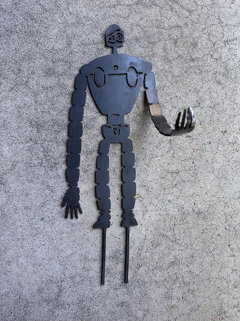 May include: A black metal robot figure with outstretched arms and legs. The robot has a detailed head, circular eyes, and segmented limbs. The figure is designed to be placed in a garden or yard as a decorative element.