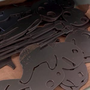 May include: A collection of dark gray metal key blanks, some with a smiling face design, are arranged in a cardboard box. The keys have a cut-out design and a keyhole. The image is taken from a slightly elevated angle.