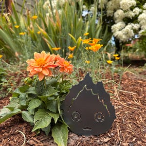 May include: A black metal decorative garden stake in the shape of a cartoon fire demon. The stake is placed in a garden bed with orange flowers and green foliage. The background features more flowers and greenery.