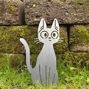 May include: A silver metal cat silhouette garden stake with large, round eyes and whiskers. The cat is sitting in a patch of green grass in front of a moss-covered stone wall. The cat's tail is curved.