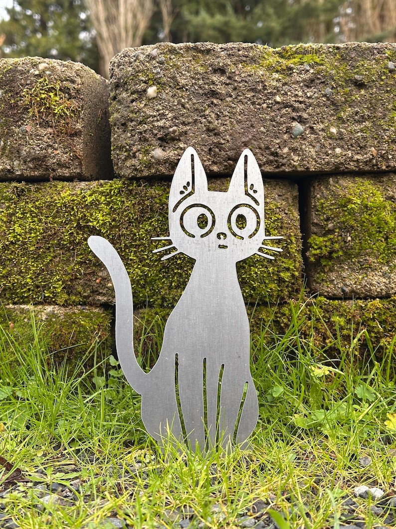 May include: A silver metal cat silhouette garden stake with large, round eyes and whiskers. The cat is sitting in a patch of green grass in front of a moss-covered stone wall. The cat's tail is curved.