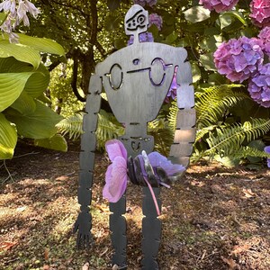 May include: A metal garden sculpture of a robot figure with a stylized design, holding a cluster of purple flowers. The robot has a dark gray finish and is positioned in a garden setting with green foliage and pink and purple hydrangeas.