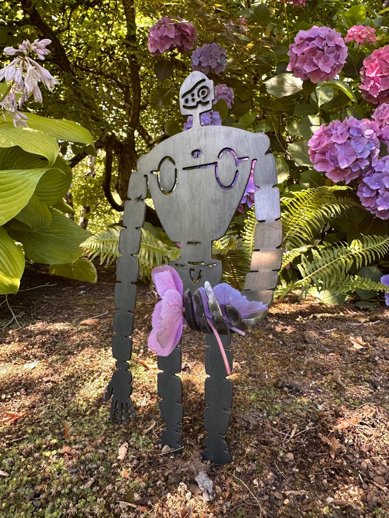 May include: A metal garden sculpture of a robot figure with a stylized design, holding a cluster of purple flowers. The robot has a dark gray finish and is positioned in a garden setting with green foliage and pink and purple hydrangeas.