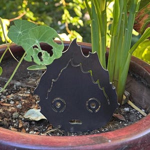 May include: A black metal garden stake in the shape of a cartoon character's face. The stake has a surprised expression with large eyes and a small mouth. It is placed in a terracotta pot with green plants and foliage in the background.