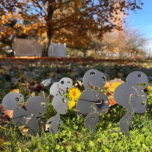 Puede incluir: Una fila de cinco esculturas de jardín de metal que representan figuras humanas estilizadas con caras y cuerpos recortados. Se encuentran en una zona cubierta de hierba, con una flor amarilla y hojas y árboles otoñales en el fondo. Diseño sencillo y moderno.