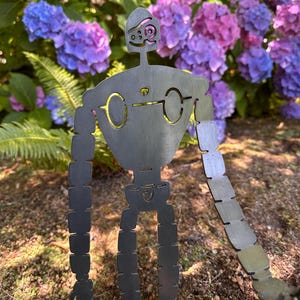 May include: A metal robot garden sculpture with a whimsical design. The robot has a dark gray finish with gold-colored accents around the eyes and chest. The sculpture is set against a backdrop of vibrant purple and blue hydrangeas.