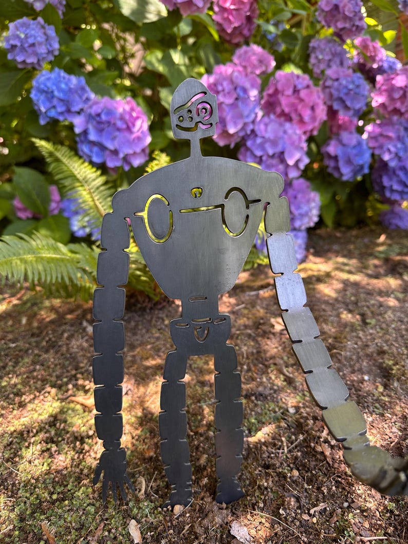 May include: A metal robot garden sculpture with a whimsical design. The robot has a dark gray finish with gold-colored accents around the eyes and chest. The sculpture is set against a backdrop of vibrant purple and blue hydrangeas.