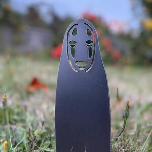 May include: A black metal garden stake featuring a stylized face design with cut-out eyes and mouth. The stake is placed in a grassy area with a blurred background of flowers and foliage. The design is inspired by the character No-Face from the animated film Spirited Away.