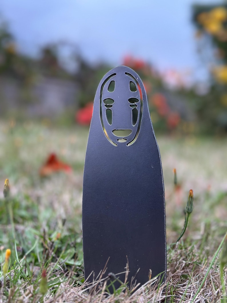 May include: A black metal garden stake featuring a stylized face design with cut-out eyes and mouth. The stake is placed in a grassy area with a blurred background of flowers and foliage. The design is inspired by the character No-Face from the animated film Spirited Away.