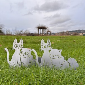 May include: Two metal cat sculptures sit in green grass. The cats are silver-colored and have large eyes and detailed features. The sculptures are placed outdoors, with a cloudy sky and a gazebo in the background.