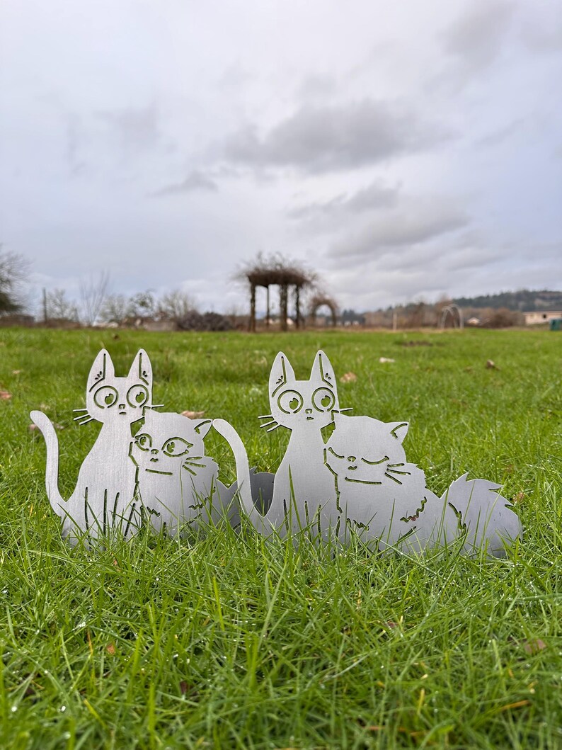 May include: Two metal cat sculptures sit in green grass. The cats are silver-colored and have large eyes and detailed features. The sculptures are placed outdoors, with a cloudy sky and a gazebo in the background.