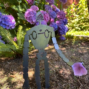 May include: A metal garden sculpture of a robot with articulated limbs, holding a purple hydrangea flower. The robot is set against a backdrop of vibrant purple and pink hydrangea blooms and green foliage.