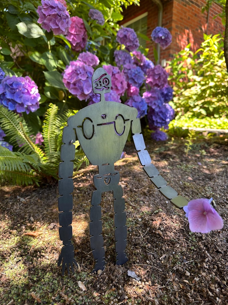 May include: A metal garden sculpture of a robot with articulated limbs, holding a purple hydrangea flower. The robot is set against a backdrop of vibrant purple and pink hydrangea blooms and green foliage.