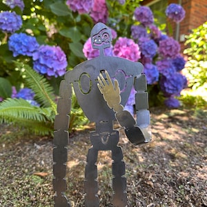 May include: A metal robot sculpture with a gold hand, set against a backdrop of vibrant purple and blue hydrangeas. The robot has a unique, stylized design with cut-out details and a textured finish, perfect for garden decor.
