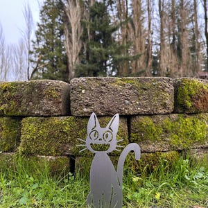 May include: A metal cat silhouette garden stake. The cat has a smiling face, pointed ears, and a curved tail. It is placed in a grassy area in front of a brick wall covered in moss. The background includes trees and a cloudy sky.