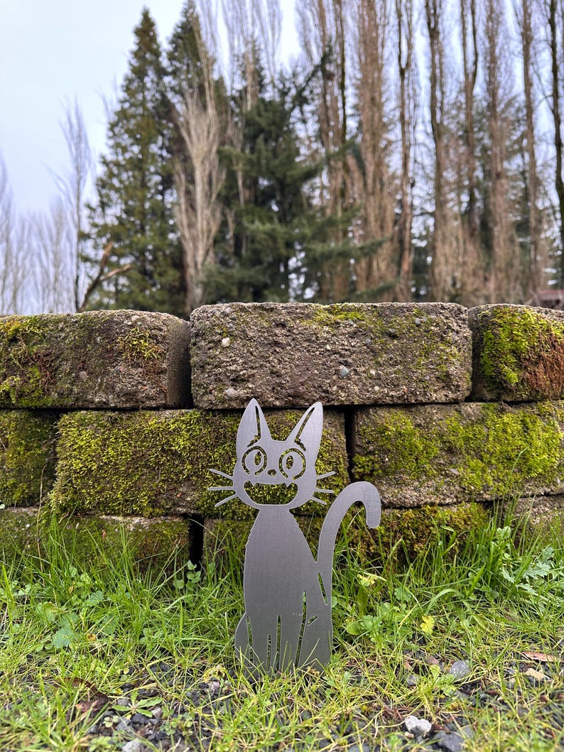 May include: A metal cat silhouette garden stake. The cat has a smiling face, pointed ears, and a curved tail. It is placed in a grassy area in front of a brick wall covered in moss. The background includes trees and a cloudy sky.