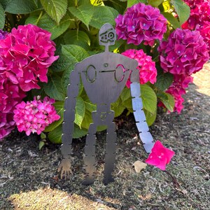 May include: A metal robot garden art piece with a stylized design, standing in front of vibrant pink hydrangea flowers. The robot has a segmented arm holding a pink flower, and is made of dark metal.